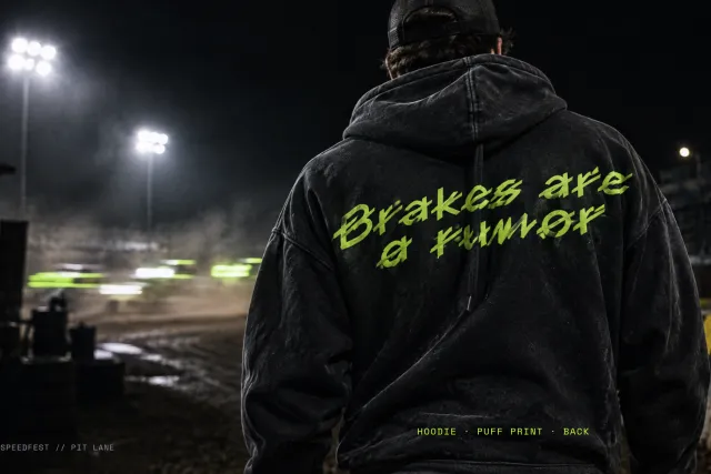 Slashrun on the back of a black hoodie, lime puff print reading Brakes are a rumor, at a floodlit dirt track