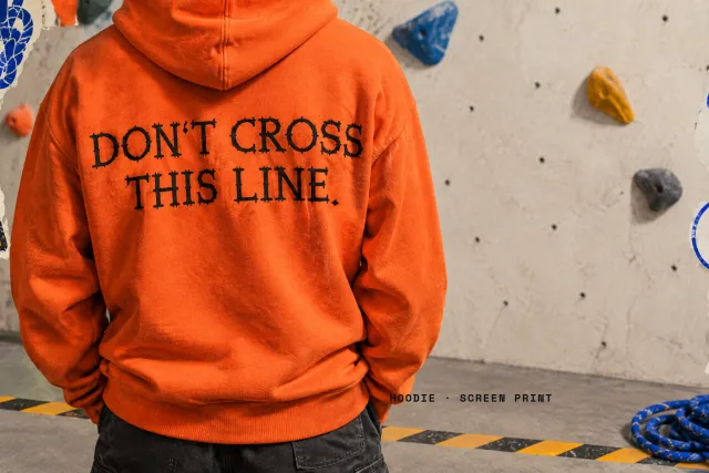 Orange hoodie with DON'T CROSS THIS LINE printed in Belay Knot, worn at a climbing wall